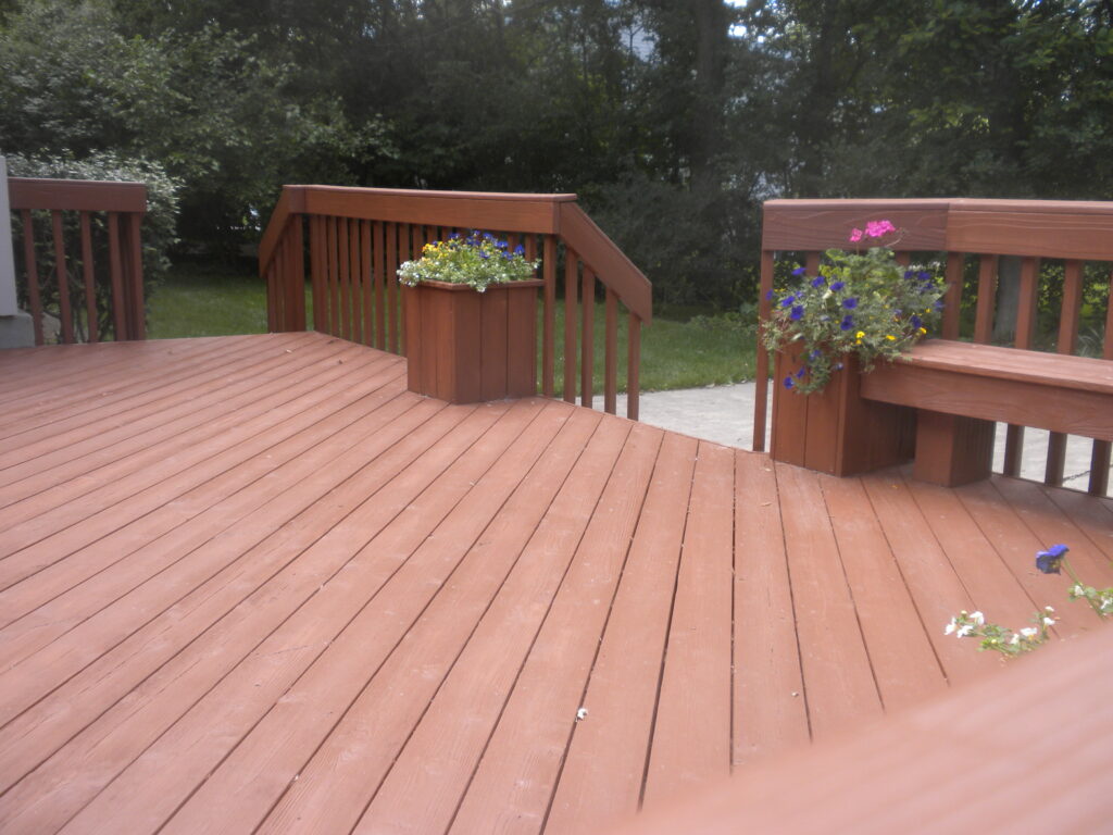 A beautiful wooden deck with matching railings and built-in planters filled with colorful flowers, overlooking a lush green backyard surrounded by mature trees.