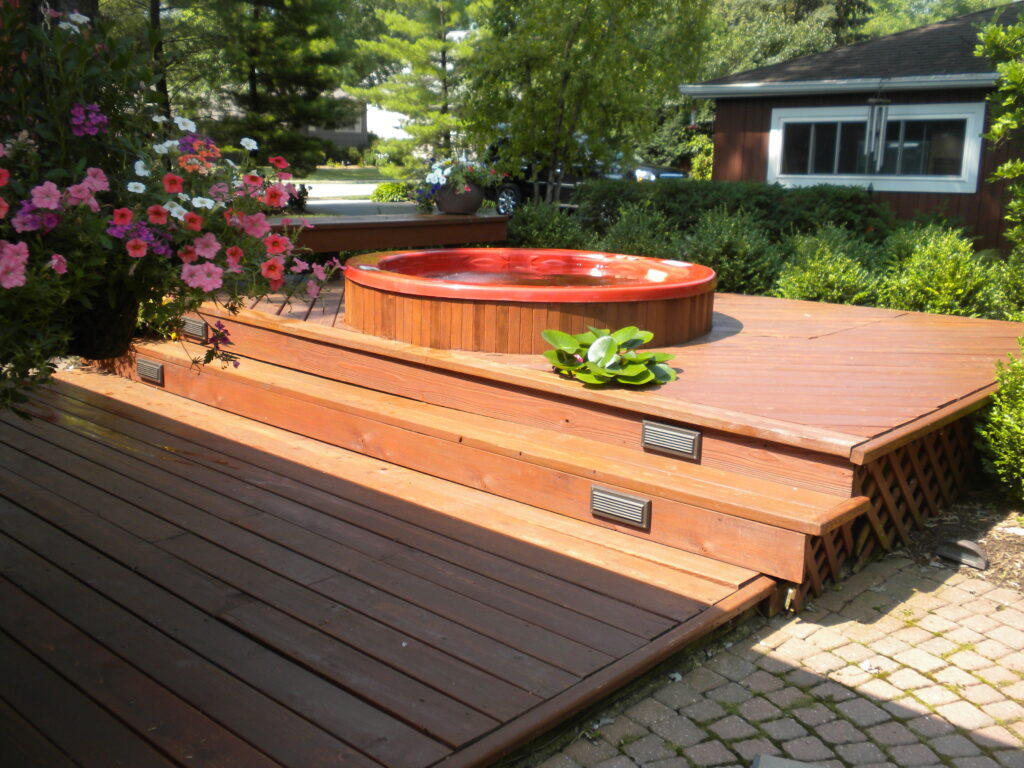 A beautiful wooden hot tub sits on a multi-level cedar deck surrounded by lush garden landscaping, colorful flowering plants, and mature trees in this peaceful backyard oasis.