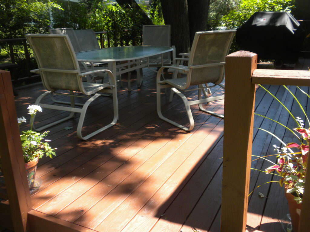 A sunny outdoor deck features modern patio furniture with curved metal chairs around a glass-top table, surrounded by lush greenery and dappled sunlight filtering through the trees above.